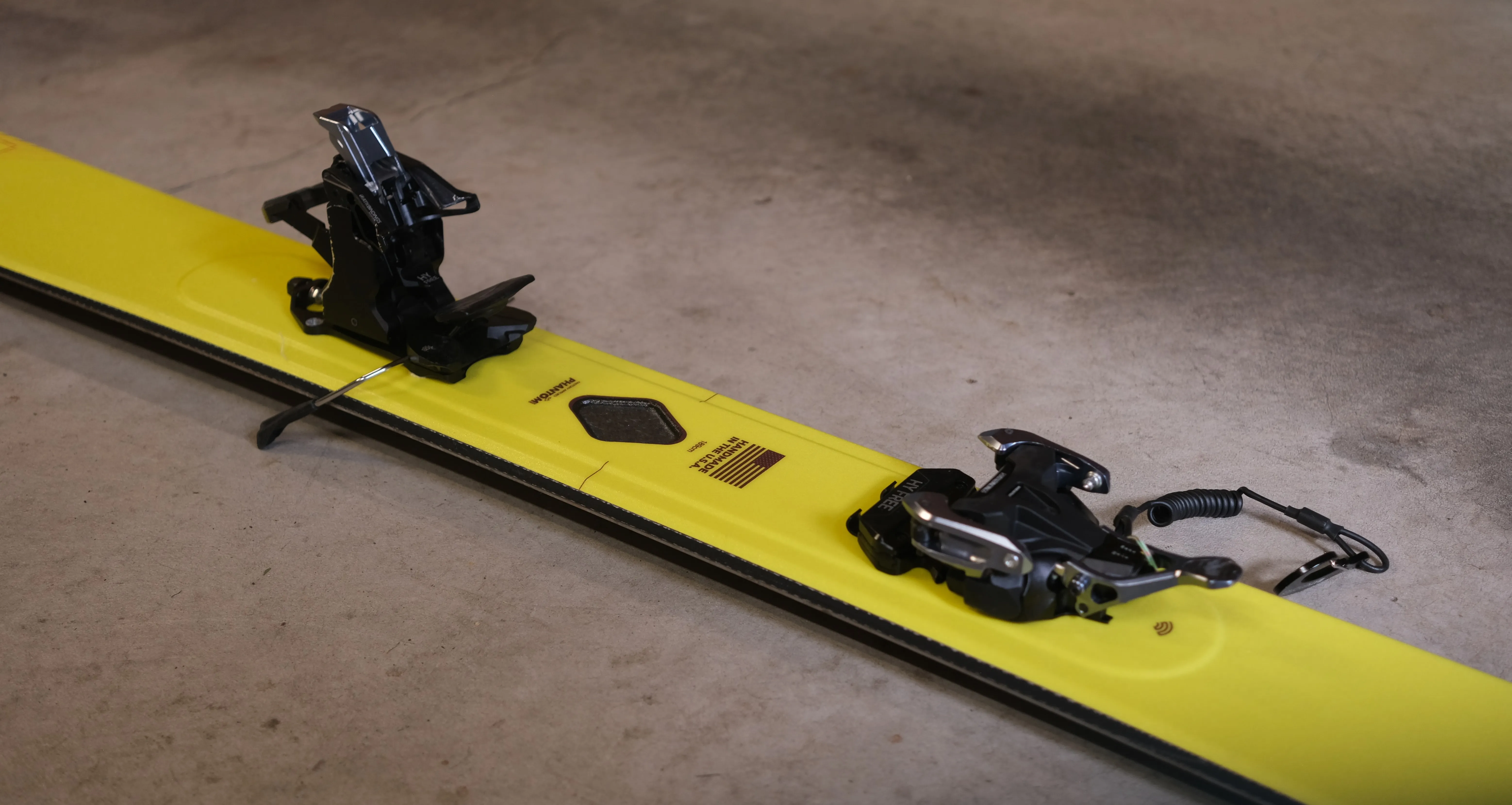 Close-up of the ATK HY Free 13 binding mounted on a yellow Phantom ski, with the toe piece on the left, the heel piece on the right, and a coiled paracord leash attached.