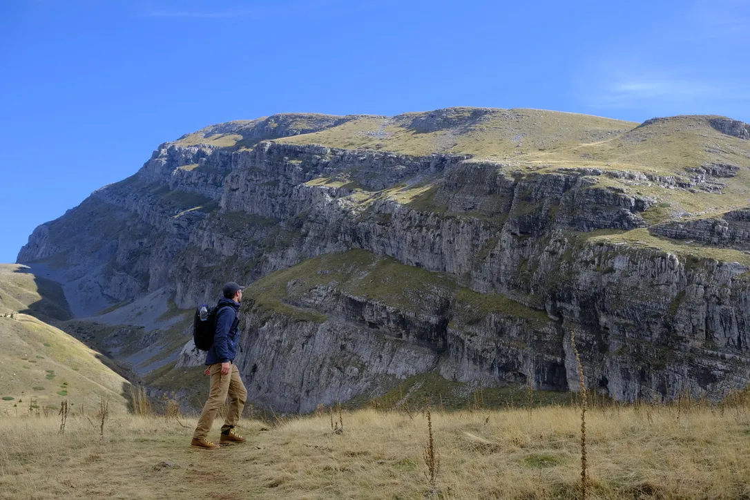 The author hiking across a grassy meadow below a dramatic limestone ridge in Greece, wearing the black Aer Travel Pack 3 Small on his back under a clear blue sky.