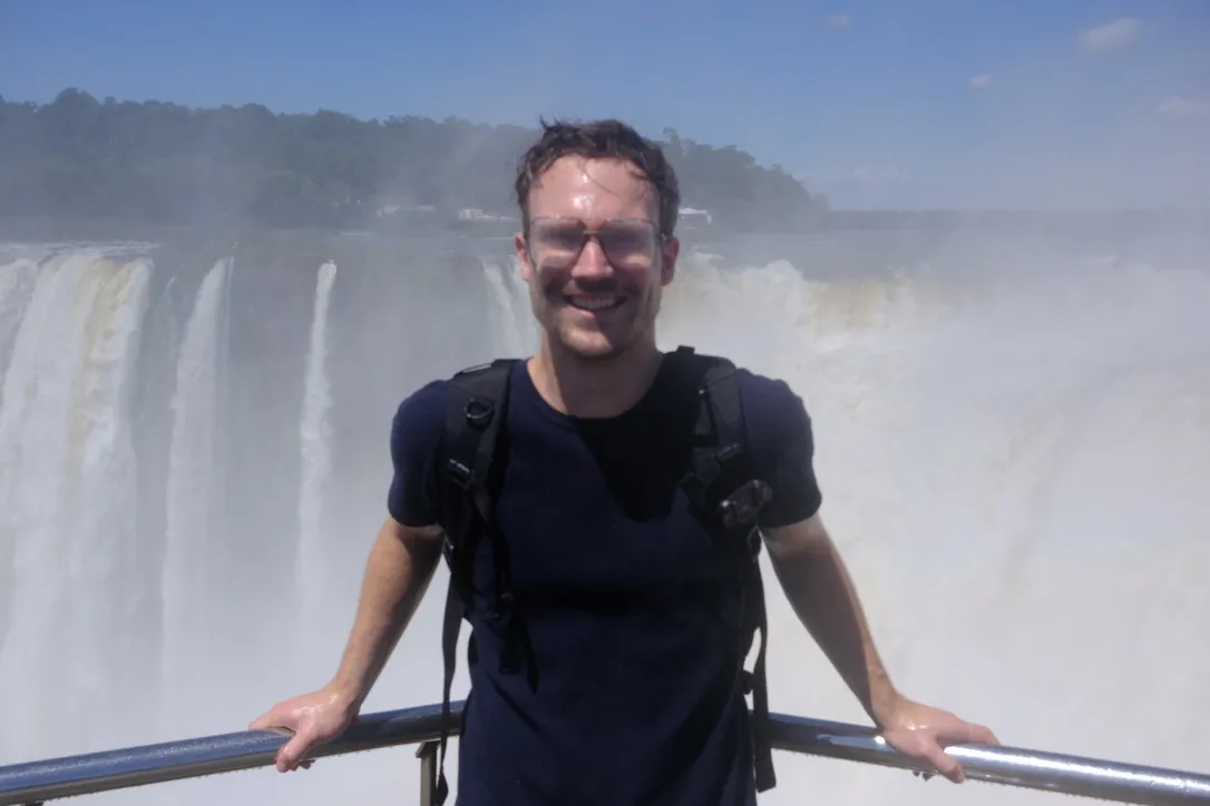 The author standing at a railing in front of the thundering, mist-shrouded cascades of Iguazu Falls in Argentina, wearing the Aer Travel Pack 3, both soaked in spray.