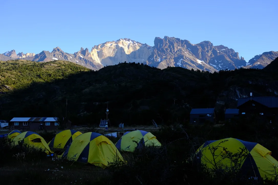 Yellow and blue premium tents lined up at a campsite with the granite peaks of the Paine Massif glowing in golden hour light behind them.
