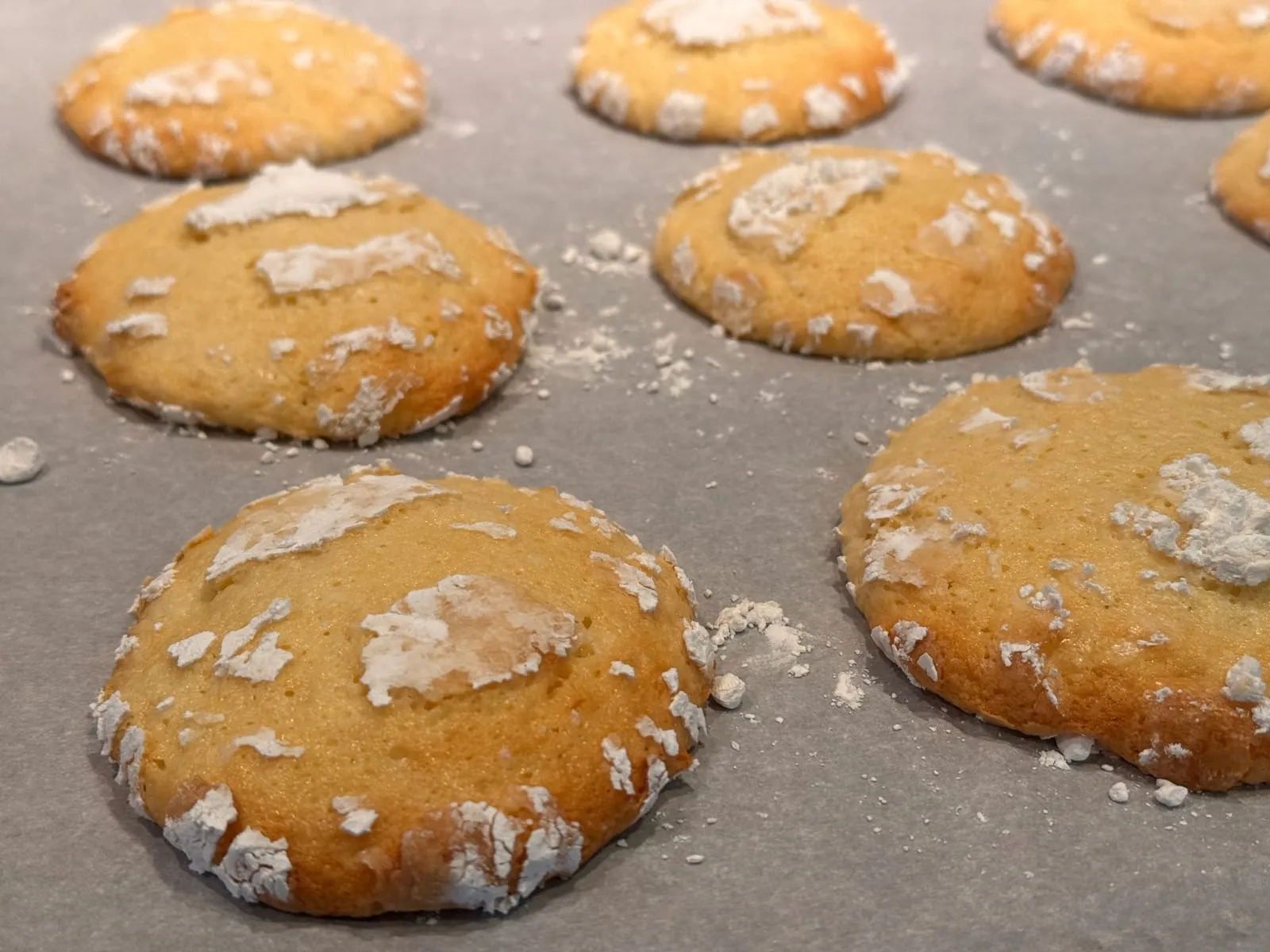 Close-up of finished lemon gooey butter cookies on parchment, golden brown edges and a cracked powdered sugar crust on top.