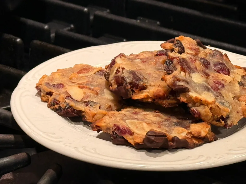 A plate of finished Marokánky cookies, lacy and golden with sliced almonds, dried cranberries, and chopped dates, dark chocolate coating their bottoms.