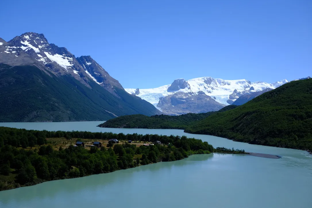 Dickson refugio nestled in green forest beside the turquoise water of Lago Dickson, with a hanging glacier and snow-covered peaks visible across the lake.