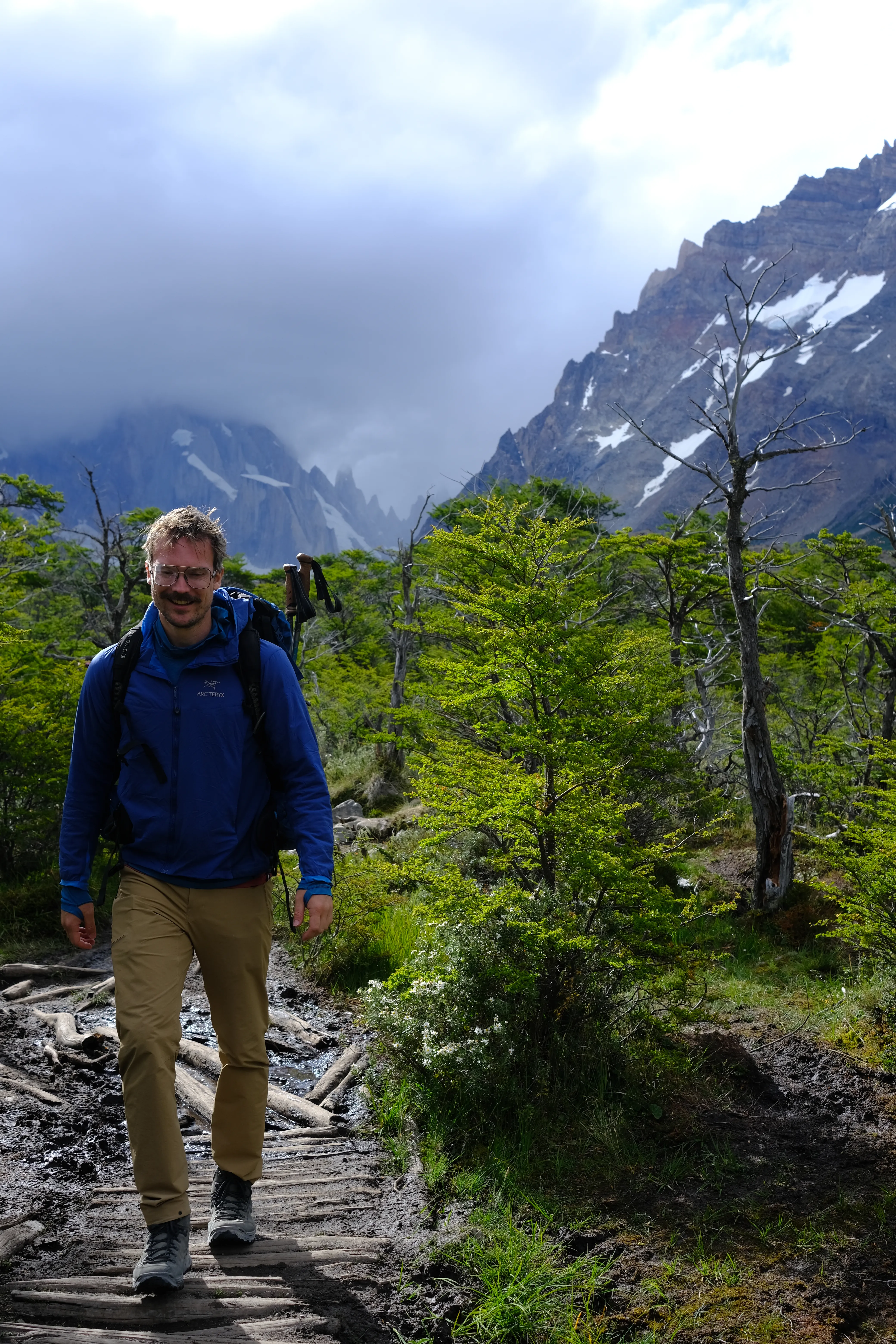 The author standing on a muddy log boardwalk on a forest trail near El Chaltén, wearing a blue jacket and Lowa Renegade boots, with green lenga forest and cloud-wrapped peaks behind him.