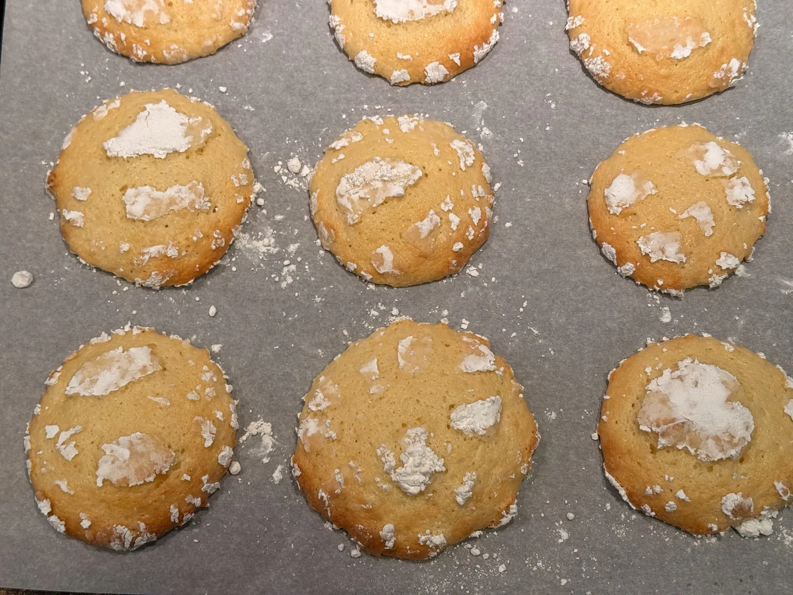 Freshly baked lemon cookies on a parchment-lined sheet, golden with cracked powdered sugar tops, straight out of the oven.