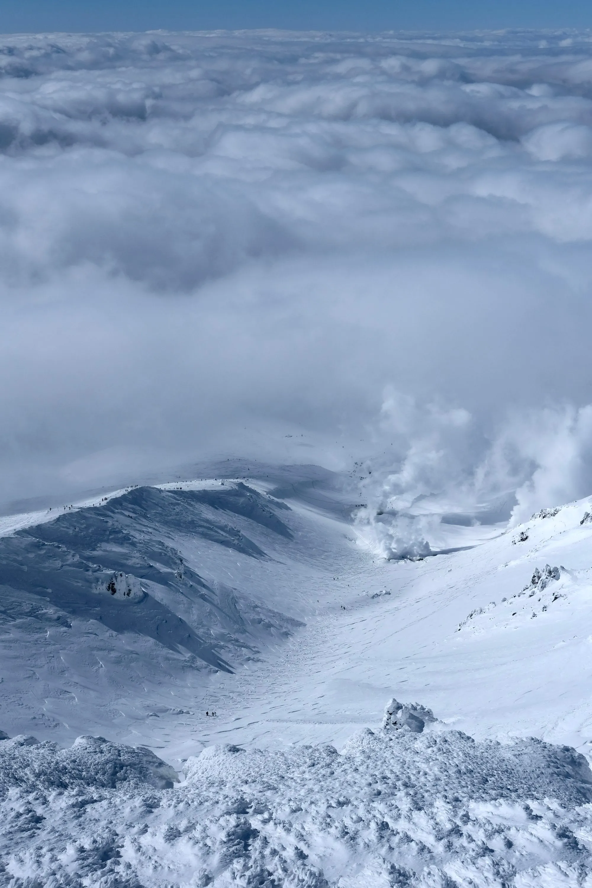 Looking down Asahidake from near the summit at fumaroles venting steam across a ridge, a thick sea of cloud filling the valley below.