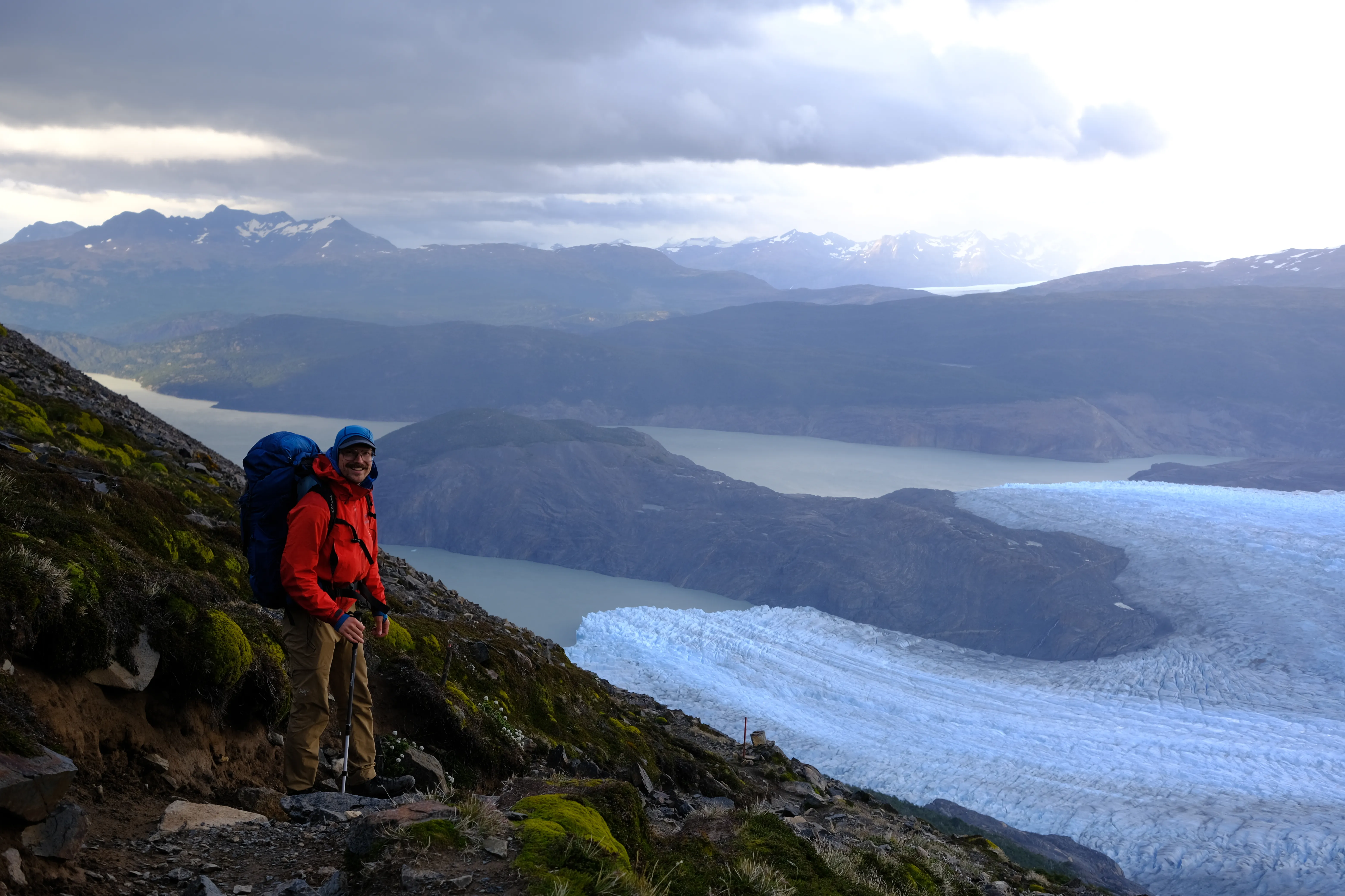 The author standing on a rocky mountainside trail high above Grey Glacier on the O Trek, wearing a red rain jacket, blue backpack, and Lowa Renegade boots, with the glacier's blue ice and Lago Grey stretching into moody clouds below.