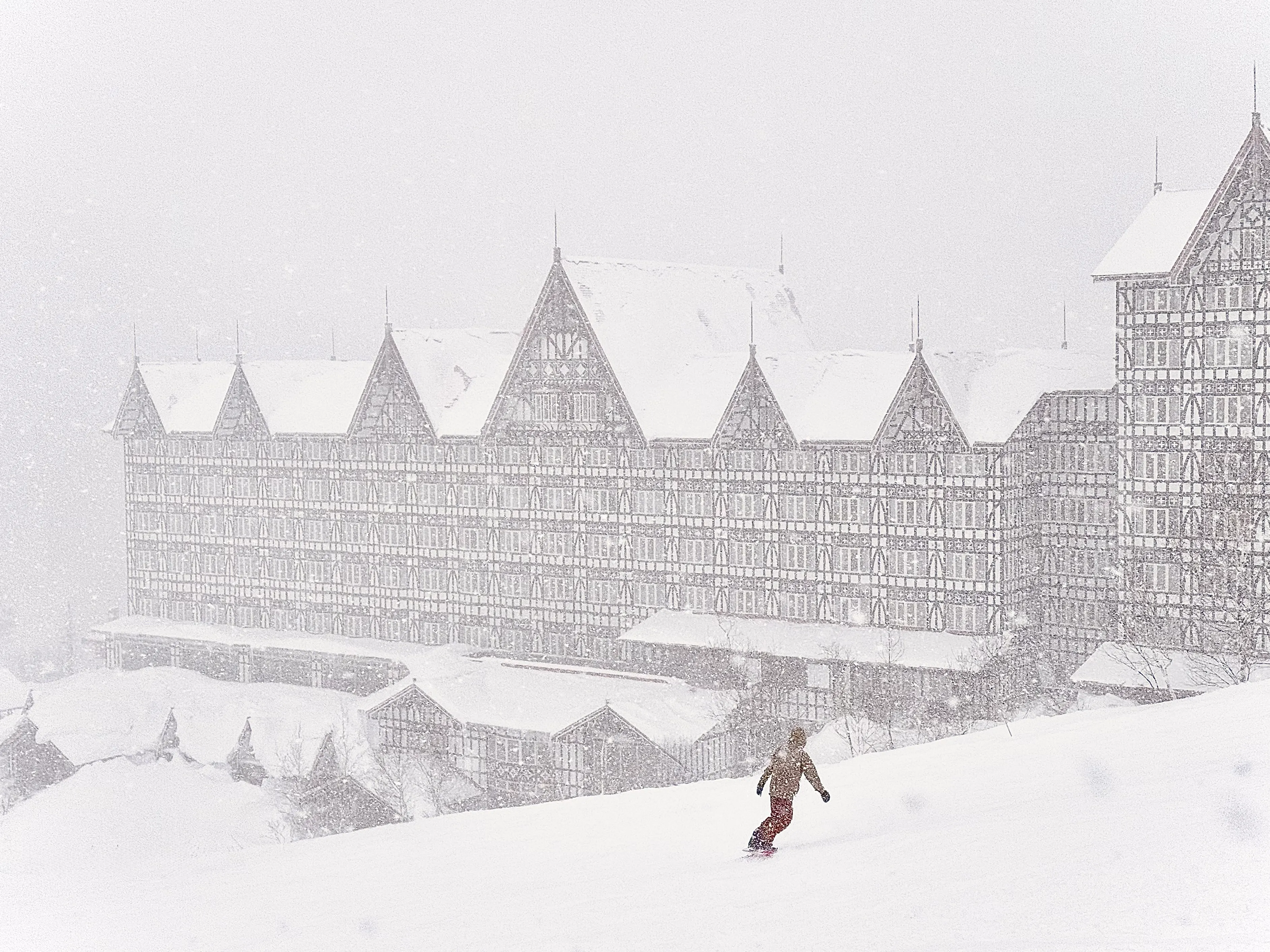 A lone skier descending a snowy slope in heavy snowfall in front of a massive half-timbered Tudor-style hotel in Hakuba, Japan, the building fading into the storm.