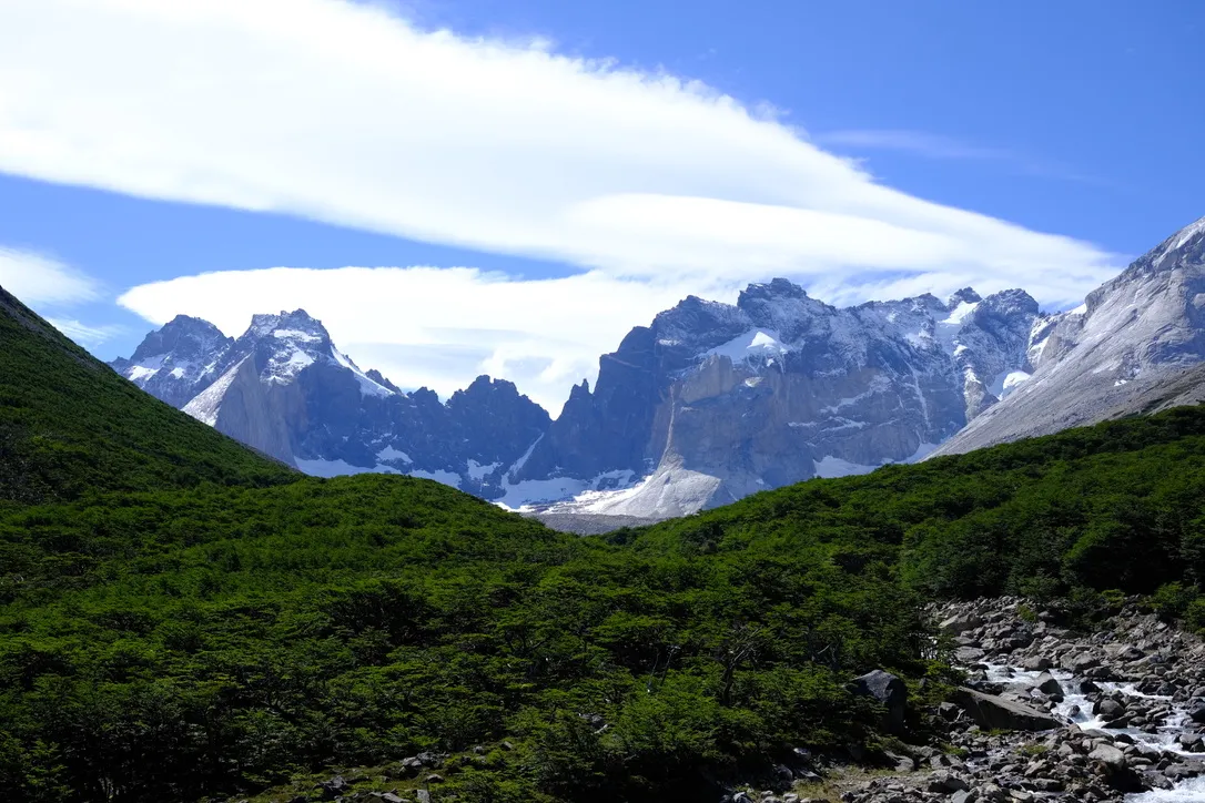 The granite walls of Valle del Frances seen from Mirador Británico, with green forested slopes leading up to dramatic snow-streaked peaks under a partly cloudy sky.