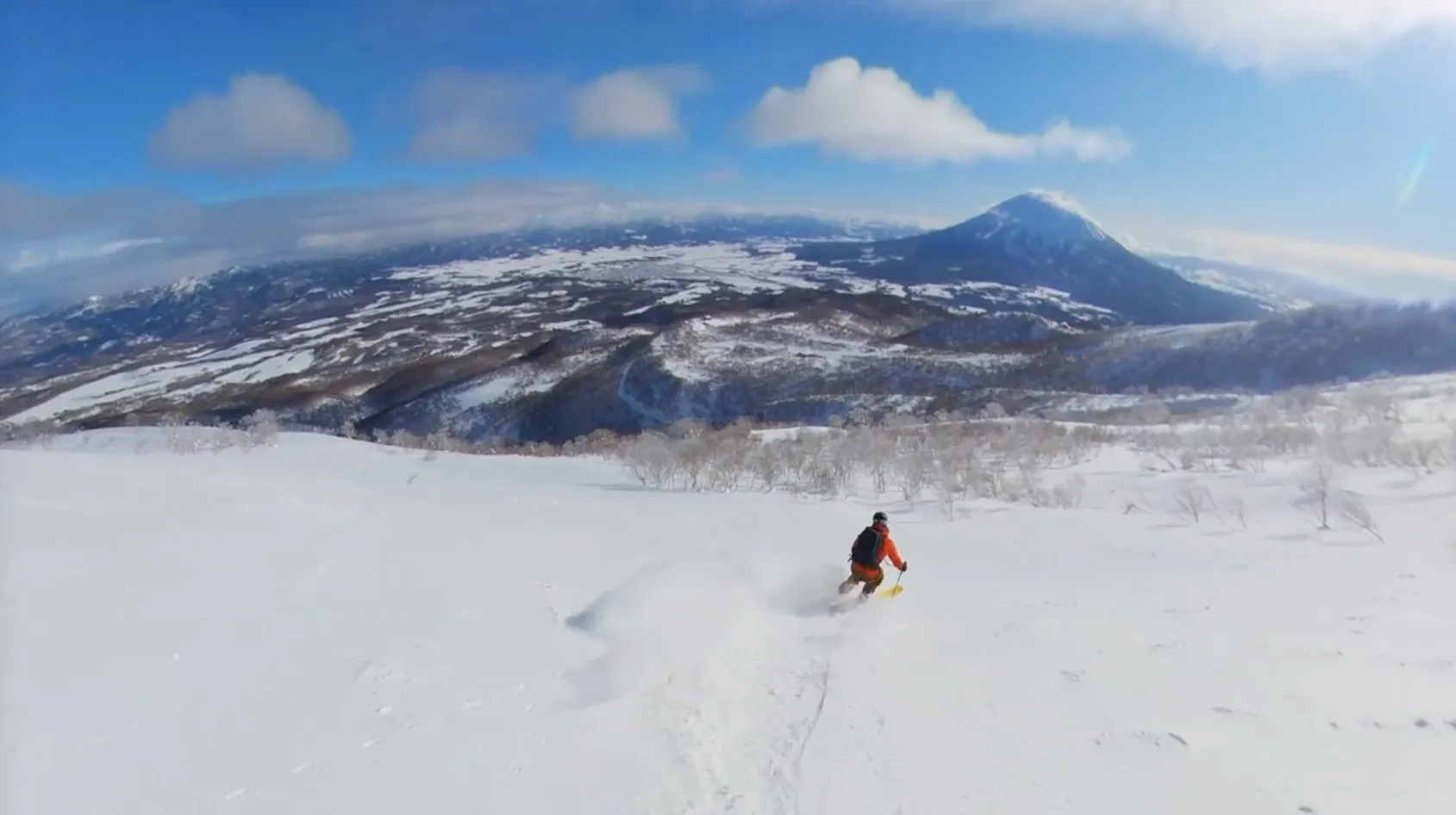 A skier descending an open, untracked powder slope toward a broad snow-covered mountain range under a blue sky with scattered clouds.