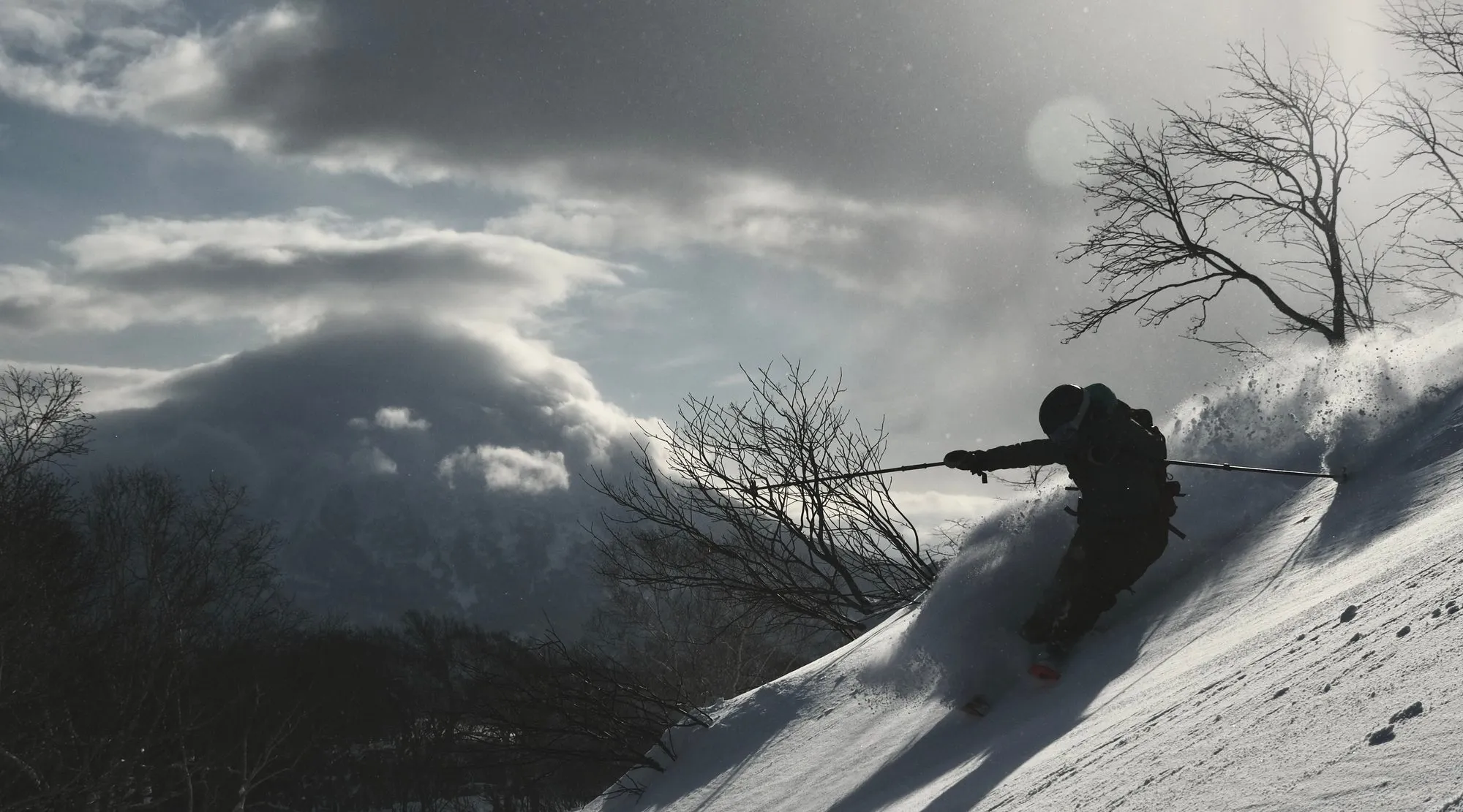 A skier carving a powder turn down a slope, snow spraying off the tails, with the cone of Mt. Yotei rising behind under a dramatic cap cloud and backlit sky.
