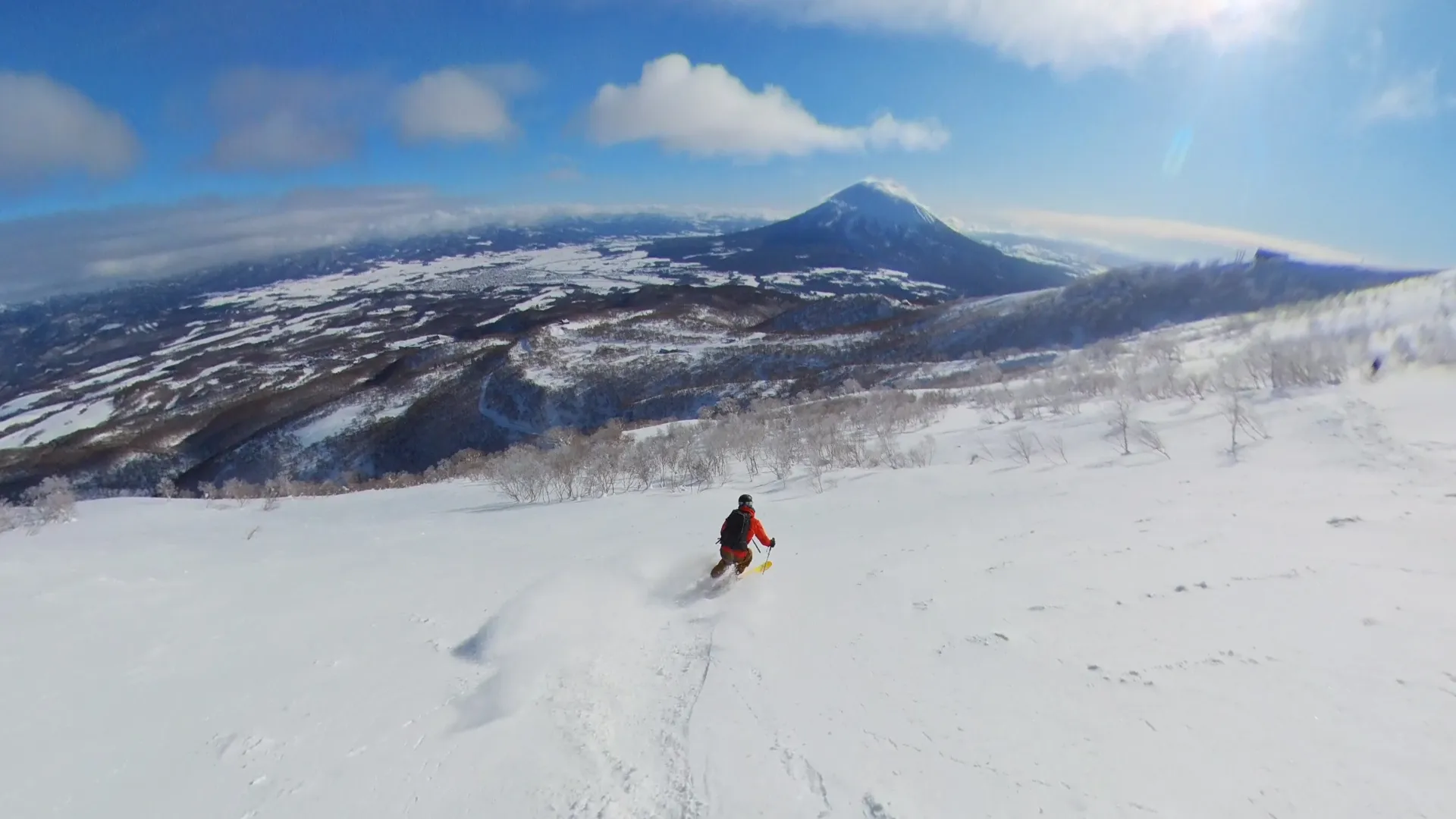 A skier in a red jacket descending an open, untracked powder slope in the Niseko sidecountry, spraying snow, with the cone of Mt. Yotei rising across the valley under a blue sky.