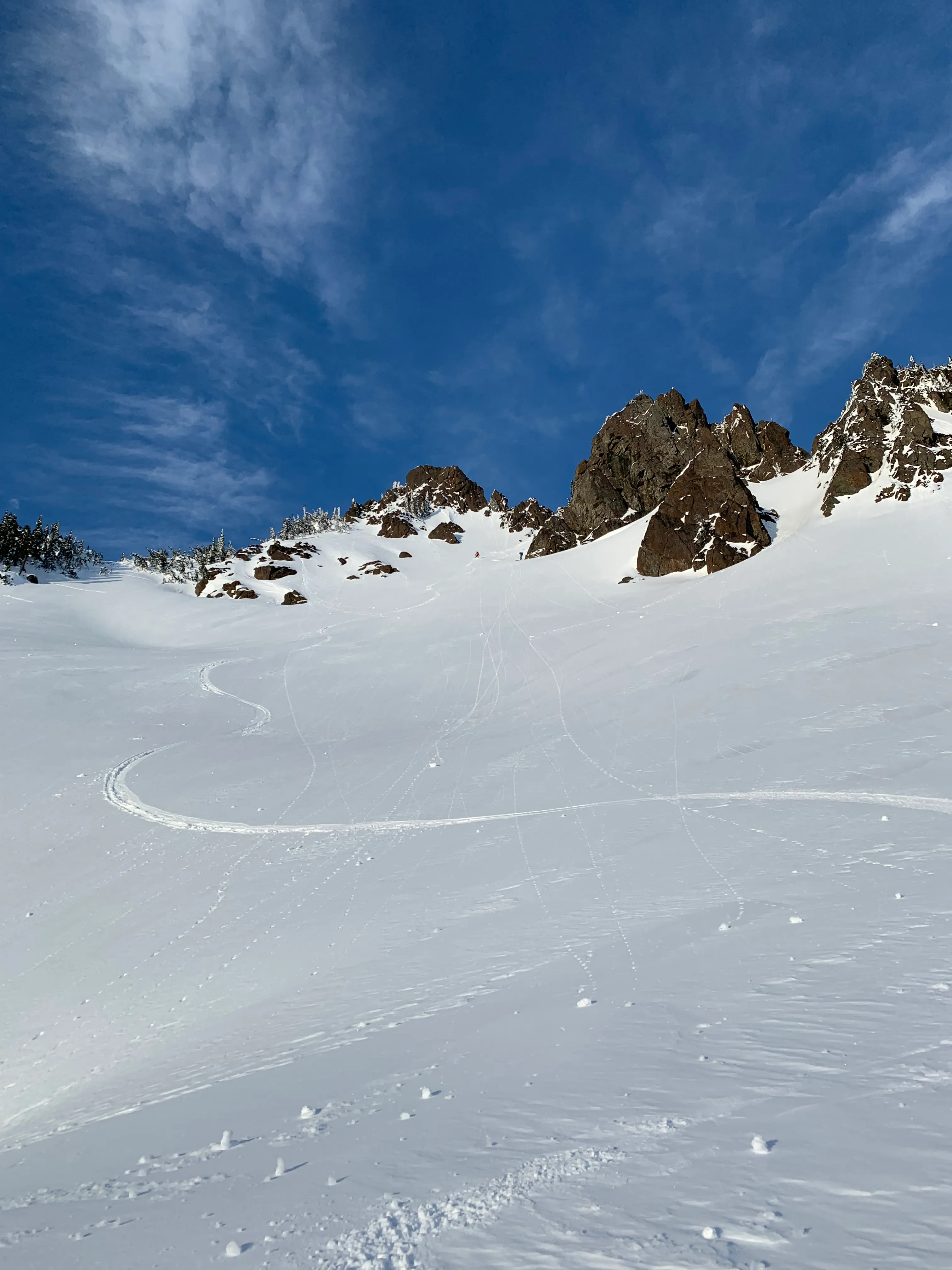 A backcountry slope in the North Cascades carved with fresh ski tracks below a jagged rocky ridge under a deep blue sky.