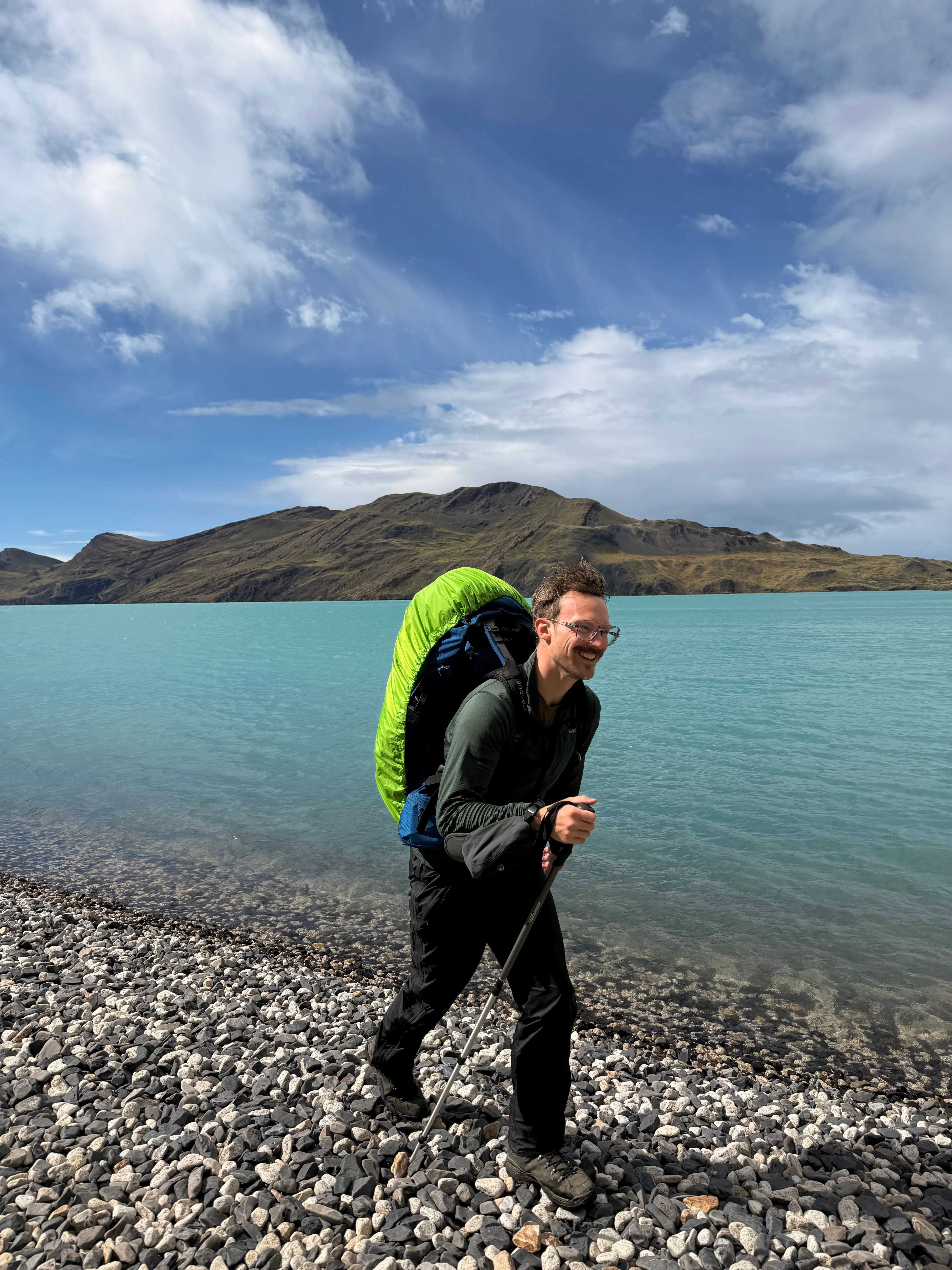 The author walking a pebble beach beside turquoise water in Torres del Paine, hair blown sideways by the wind, wearing black rain pants, a pack with a bright green rain cover, and Lowa Renegade boots on the stones.