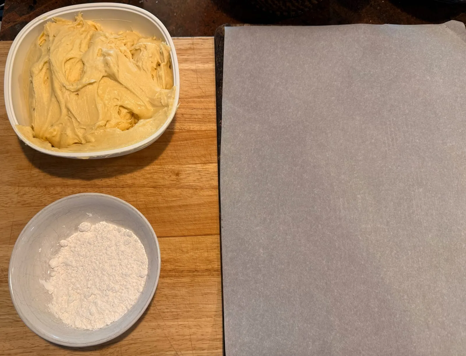 A tub of pale yellow cookie dough and a small bowl of powdered sugar on a wooden board, next to a parchment-lined baking sheet.