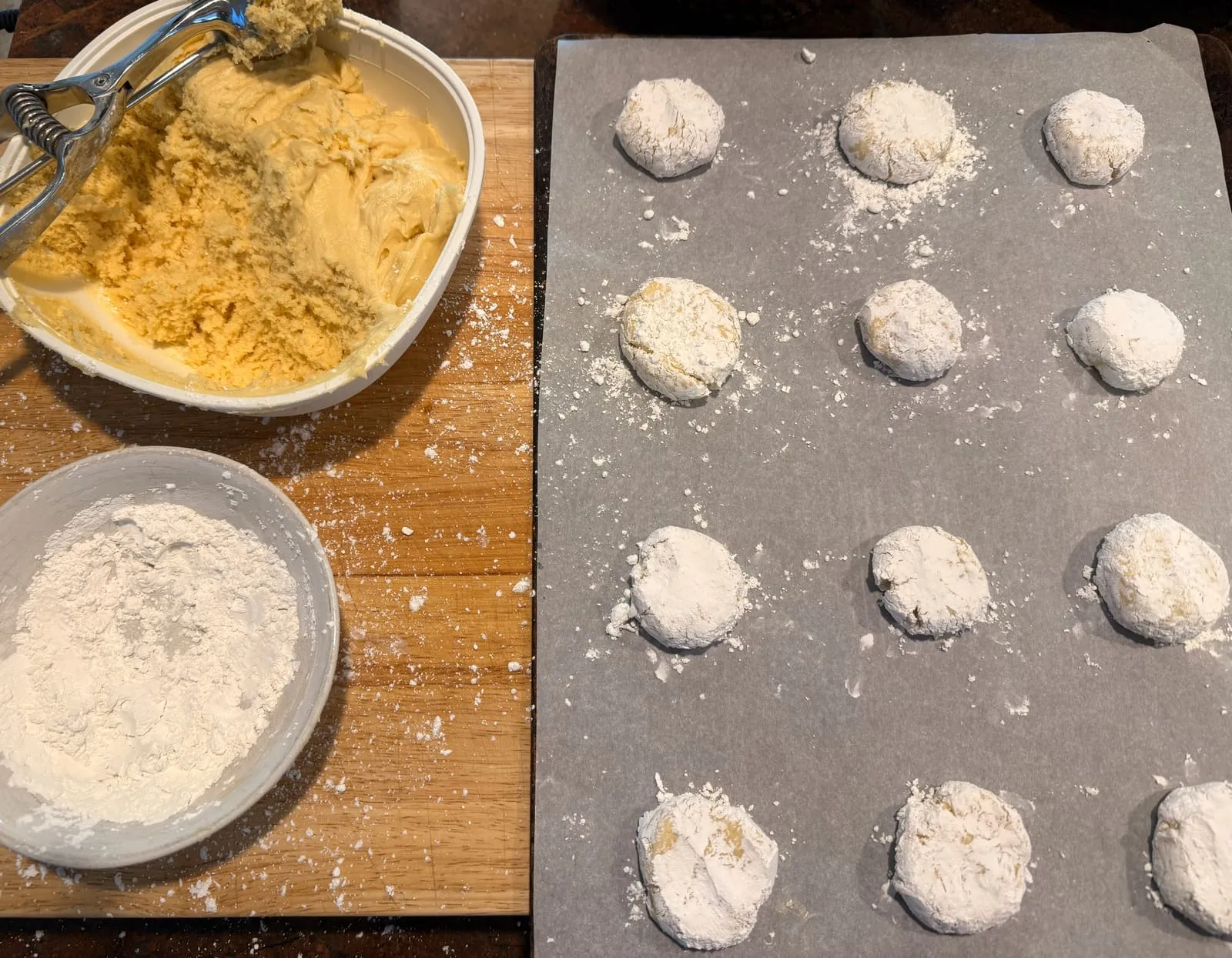 Powdered-sugar-coated dough balls spaced out in rows on a parchment-lined baking sheet, with the dough and sugar bowls alongside.