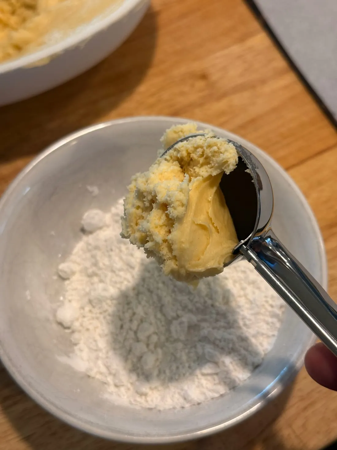 An ice cream scoop of cookie dough held over a bowl of powdered sugar.