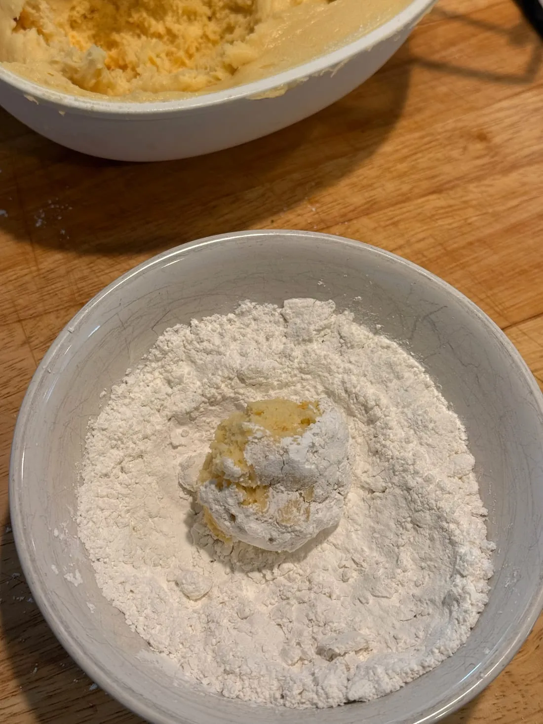 A ball of cookie dough sitting in a bowl of powdered sugar, half coated in white.