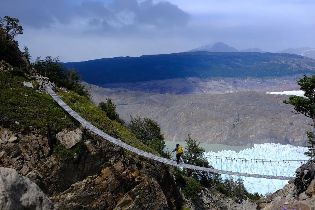 A hiker with a yellow pack crossing a long, narrow wooden suspension bridge above a steep canyon, with the white ice of Grey Glacier filling the valley behind.