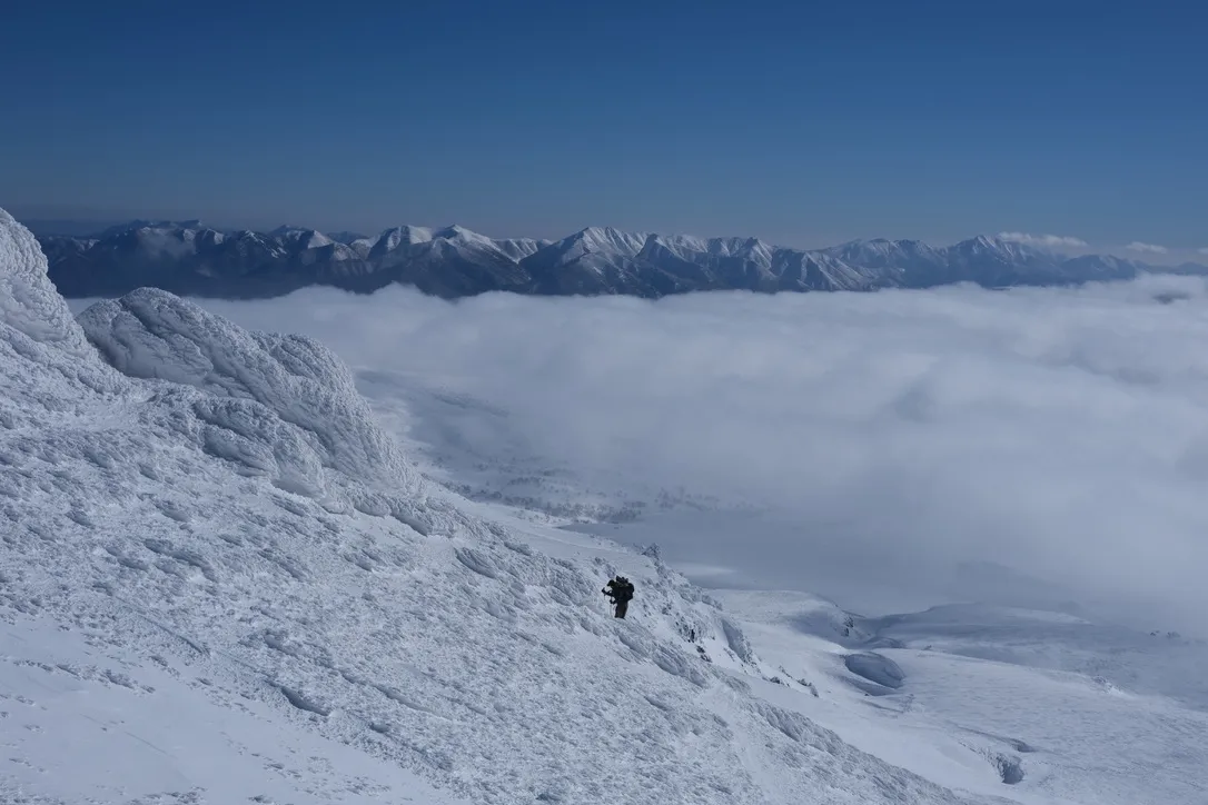 A lone skier with a large pack climbing a steep, rime-iced ridge above a thick sea of clouds, a long range of snow-covered peaks on the horizon under a deep blue sky.