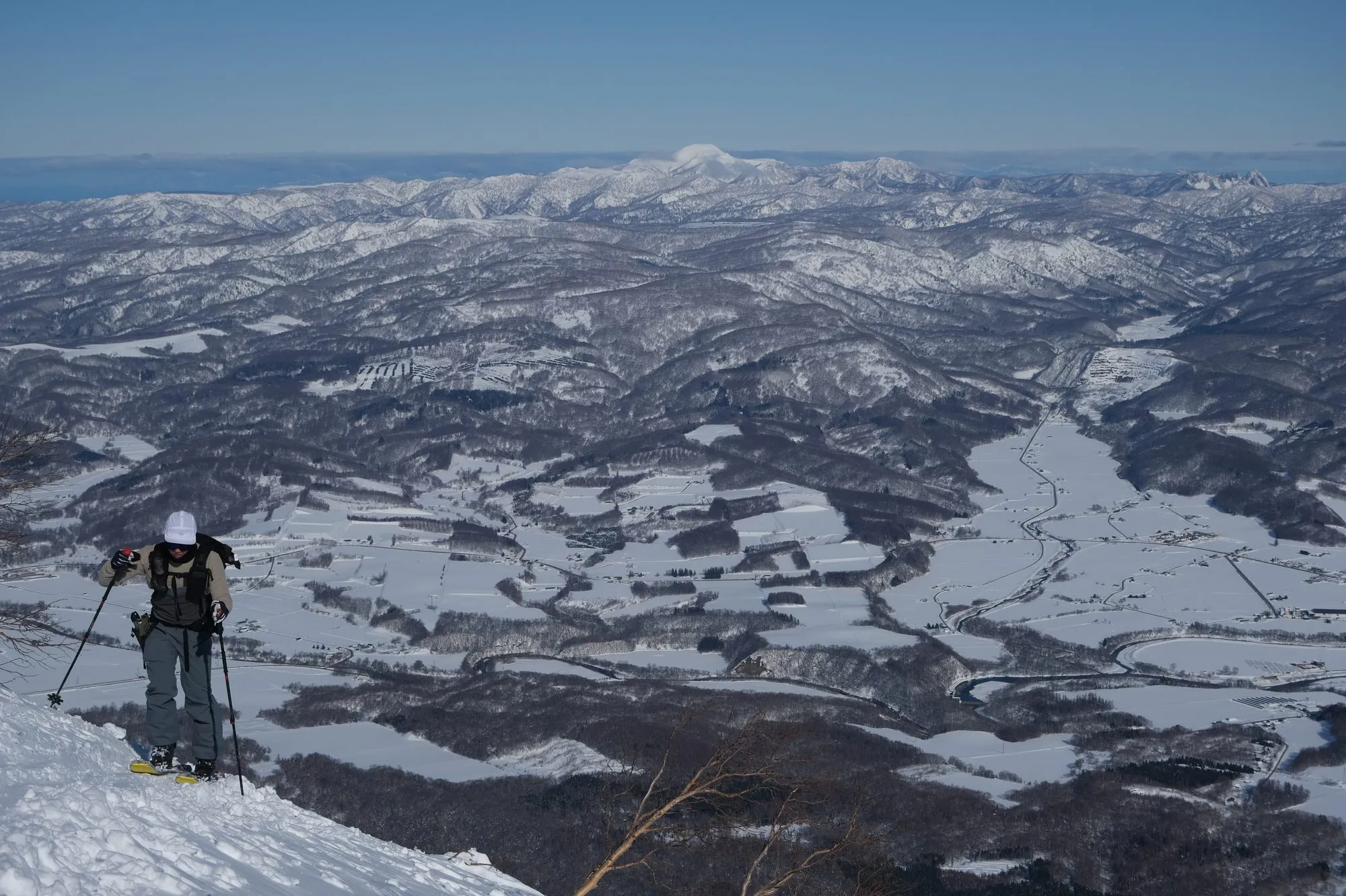 A lone skier skinning up a steep ridge on Mt. Yotei, the vast snowy Niseko farmland and a distant volcano far below.