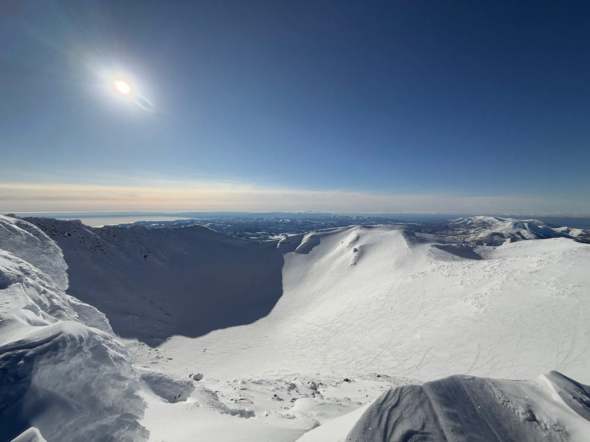 The wide, snow-filled summit crater of Mt. Yotei seen from the rim, sun flaring in a deep blue sky with the distant Sea of Japan on the horizon.