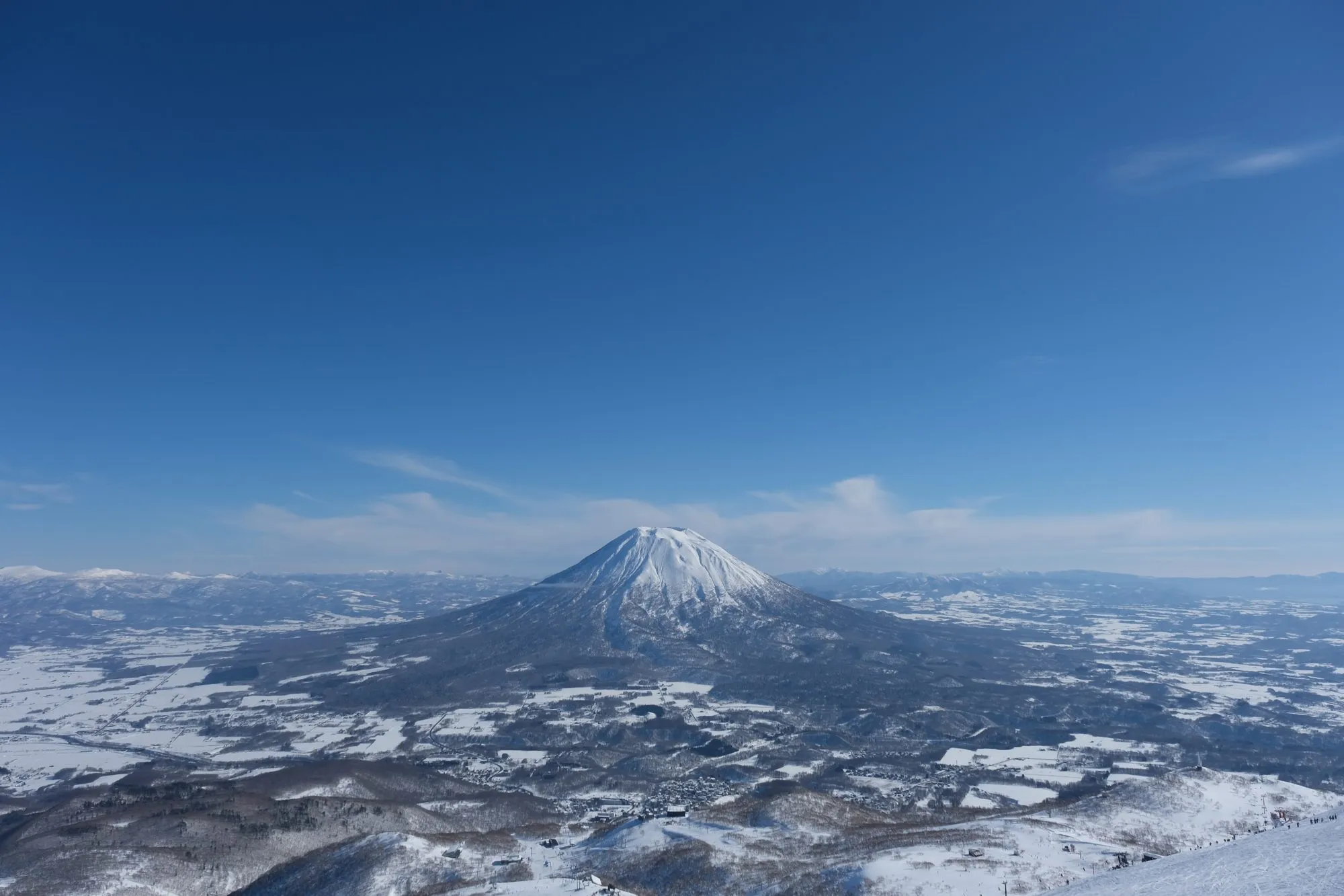 The symmetrical snow-covered cone of Mt. Yotei rising over the patchwork of snowy farmland in the Niseko valley under a clear blue sky.
