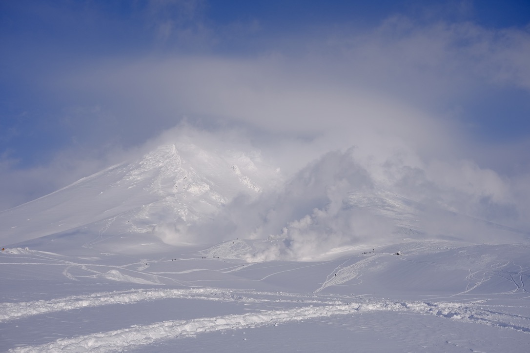 Snow-covered Asahidake volcano with volcanic smoke billowing from the summit, ski tracks visible in the foreground powder, and tiny figures of skiers dotting the slopes below.
